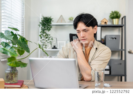 An overworked Asian businessman sits at his desk, massaging his shoulder and feeling uncomfortable. An overworked Asian businessman sits at his desk, massaging his shoulder and feeling uncomfortable. 122552792