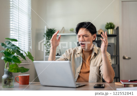 Angry Asian businessman sits at his desk, looking at his laptop screen with a frustrated expression. 122552841