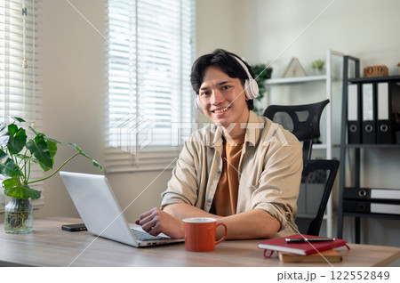 A creative Asian businessman is sitting in front of a laptop in his office, smiling at the camera. 122552849