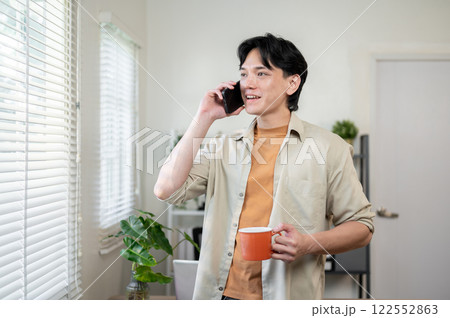 A positive Asian businessman stands in his office with a coffee cup in hand, talking on the phone. 122552863