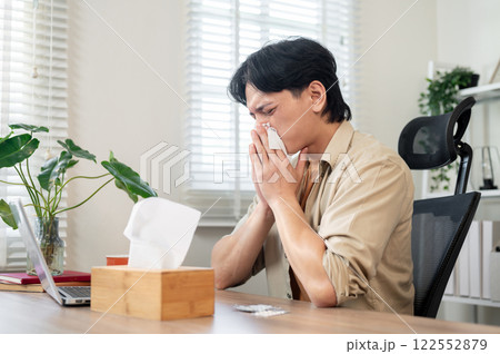 An unwell Asian businessman sits at his desk in the office, blowing his nose while working. An unwell Asian businessman sits at his desk in the office, blowing his nose while working. 122552879