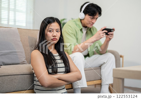 An unhappy girl sits in the living room, feeling ignored by her boyfriend as he plays a mobile game. 122553124
