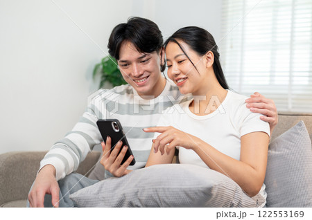 A happy young Asian couple sits together on a sofa in the living room, watching a video on a phone. A happy young Asian couple sits together on a sofa in the living room, watching a video on a phone. 122553169