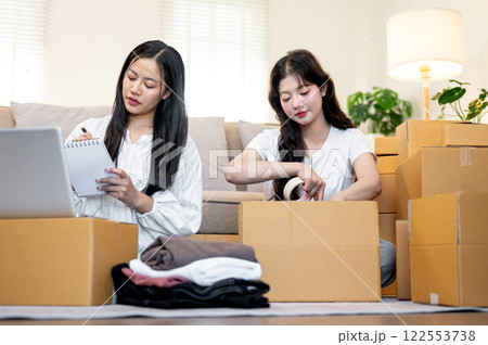 Two Asian female online shop owners are packing goodies into cardboard boxes, preparing items. Two Asian female online shop owners are packing goodies into cardboard boxes, preparing items. 122553738