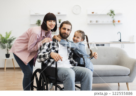 Portrait of Caucasian family with father in wheelchair, mother, and young daughter in kitchen. Warm and supportive family bond captured in comfortable, modern home environment. Portrait of Caucasian family with father in wheelchair, mother, and young daughter in kitchen. Warm and supportive family bond captured in comfortable, modern home environment. 122555886