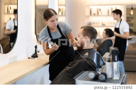 Young woman barbershop employee stands next to male client, discussing details of haircut Young woman barbershop employee stands next to male client, discussing details of haircut 122555892