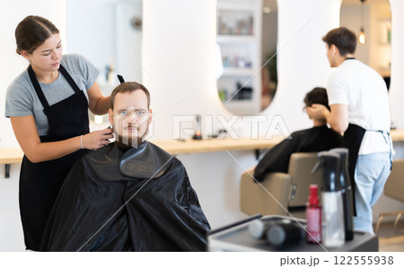 Young woman barbershop employee stands next to male client, discussing details of haircut 122555938