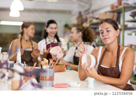 Young woman sits near table and decorates clay ceramic craft vase, plate 122556056
