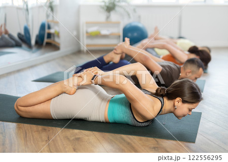 Happy young woman with sports people doing bow pose of yoga on mat together in yoga studio Happy young woman with sports people doing bow pose of yoga on mat together in yoga studio 122556295