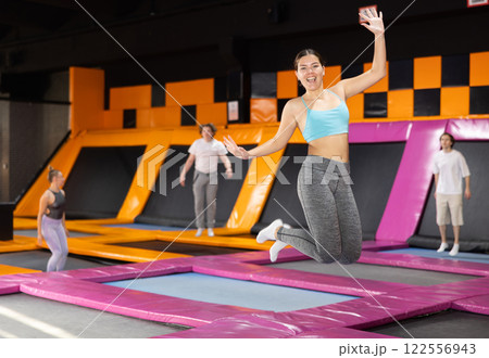 Excited young woman in blue top and gray leggings having great time while jumping on colorful trampoline in game club 122556943