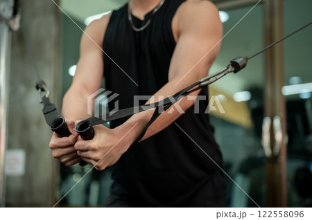 A close-up of a muscular Asian man working out on a cable machine in the gym, pulling the cables. 122558096