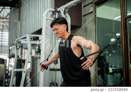A serious Asian male athlete is using a cable machine in the gym to train his biceps and shoulders. 122558098