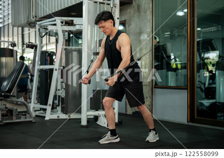 A serious, muscular Asian male athlete is using a cable machine in the gym. A serious, muscular Asian male athlete is using a cable machine in the gym. 122558099
