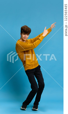 Full-length portrait of joyful teen boy in casual clothes yellow sweater and black jeans, cheerfully dancing against blue studio background Full-length portrait of joyful teen boy in casual clothes yellow sweater and black jeans, cheerfully dancing against blue studio background 122565485