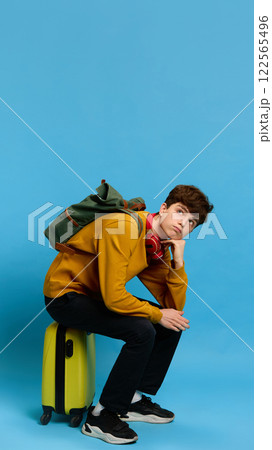 Bored and tired teen boy in casual wear sitting on suitcase with backpack, waiting for his trip against blue studio background Bored and tired teen boy in casual wear sitting on suitcase with backpack, waiting for his trip against blue studio background 122565496