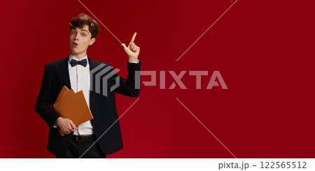 Portrait of young man, student on formal wear with bow tie standing with papers and raising finger up, showing gesture of idea against red studio background Portrait of young man, student on formal wear with bow tie standing with papers and raising finger up, showing gesture of idea against red studio background 122565512