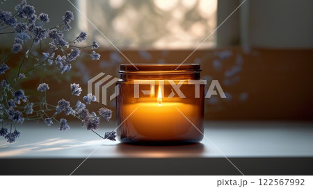 Amber jar candle glowing softly on a windowsill, surrounded by lavender flowers in sunlight 122567992