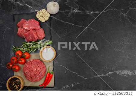 Top view of minced beef patty lying on paper surrounded by seasonings, red hot pepper and tomato on a branch, next to cut meat and garlic separated by a sprig of rosemary on a dark background with 122569013