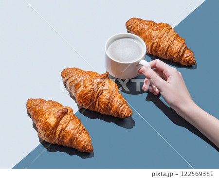Hand holding cup of cappuccino or latte coffee on blue background with fresh croissants and shadow. Hand holding cup of cappuccino or latte coffee on blue background with fresh croissants and shadow. 122569381
