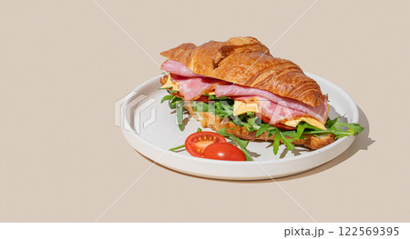 Croissant sandwich with ham, cheese and arugula on a plate on a yellow background with a shadow. Croissant sandwich with ham, cheese and arugula on a plate on a yellow background with a shadow. 122569395