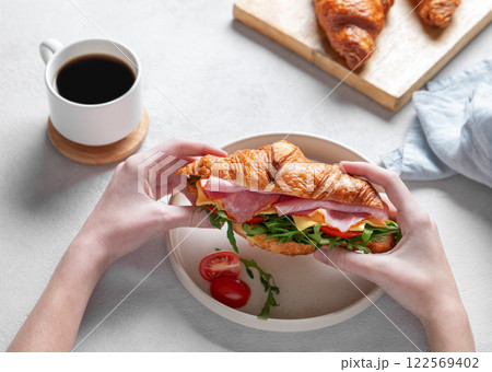 Hands holding fresh croissant sandwich with ham, cheese, tomato and arugula on a plate Hands holding fresh croissant sandwich with ham, cheese, tomato and arugula on a plate 122569402