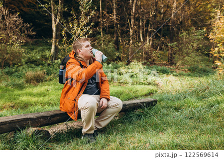 Happy blonde man with backpack having a halt after hiking. 30s Hiker drinking water from water bottle or hot drink from blue thermos. Active lifestyle. 122569614