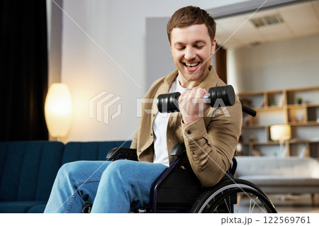 Physical rehabilitation for disabled people. Happy guy in wheelchair making exercises with dumbbell at home. Cheerful impaired young man working out with weights indoors Physical rehabilitation for disabled people. Happy guy in wheelchair making exercises with dumbbell at home. Cheerful impaired young man working out with weights indoors 122569761
