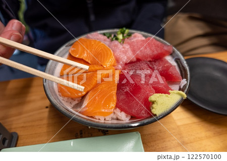 Person is enjoying a bowl of sushi using chopsticks. Salmon. Wasabi. Tuna. Rice Person is enjoying a bowl of sushi using chopsticks. Salmon. Wasabi. Tuna. Rice 122570100
