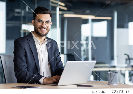 Portrait of successful satisfied businessman, man in business suit looking at camera. Joyful boss at workplace inside office using laptop. 122570158