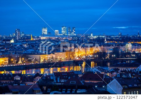 The National Theatre stands illuminated against the evening sky in Prague, with modern buildings of Pankrac rising in the background. Reflections shimmer in the water. 122571047