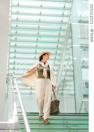 Wide shot of a Caucasian pretty young elegant woman in a straw hat and carrying a bag descending transparent glass steps. Vertical. In the background is a modern business building 122572320