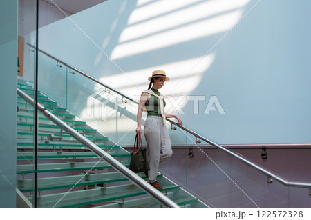 Wide view of a Caucasian pretty young elegant woman in a straw hat and carrying a bag descending transparent glass steps. Sunbeams at the wall. Copy space Wide view of a Caucasian pretty young elegant woman in a straw hat and carrying a bag descending transparent glass steps. Sunbeams at the wall. Copy space 122572328