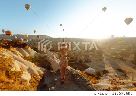 Happy girl in hat stands on mountain with flying air balloons on background. View from the back. Happy girl in hat stands on mountain with flying air balloons on background. View from the back. 122572690