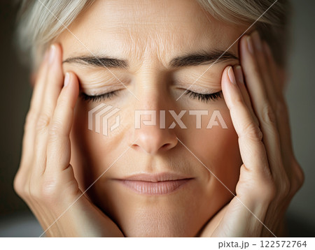 A close-up of a middle-aged woman with closed eyes holding her temples 122572764