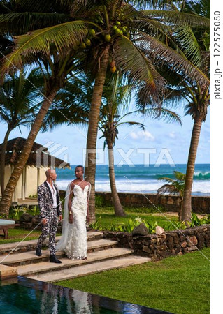 Ambiguous couple at tropical villa wedding. Black person in white dress, partner in bold suit walk by ocean. LGBTQ-themed event, diversity celebrated. Scenic, inclusive celebration. 122575080