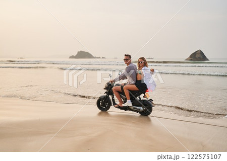 Couple rides motorbike on sandy beach at sunset. Man drives scooter with woman enjoying view. Travel fun, explore coast on budget vacation. Adventure lovers embrace leisure ocean journey together. Couple rides motorbike on sandy beach at sunset. Man drives scooter with woman enjoying view. Travel fun, explore coast on budget vacation. Adventure lovers embrace leisure ocean journey together. 122575107