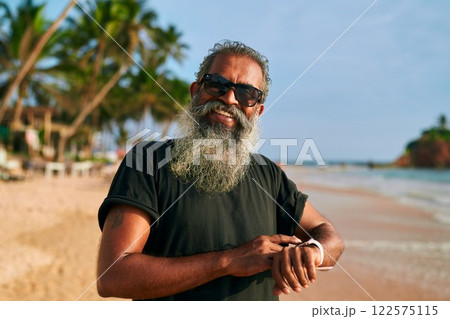 Stylish gray-bearded senior with sunglasses uses smart watch on tropical beach. Modern tech-savvy grandpa enjoys active lifestyle and sunny day. Elderly man embraces digital fitness. Stylish gray-bearded senior with sunglasses uses smart watch on tropical beach. Modern tech-savvy grandpa enjoys active lifestyle and sunny day. Elderly man embraces digital fitness. 122575115