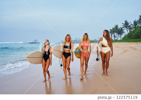 Young women walk with surfboards on sandy beach. Friends enjoy summer vacation. Surfing, tropical ocean. Active lifestyle. Palm trees, waves, sunset sky. Outdoor leisure, coastal exploration. 122575119