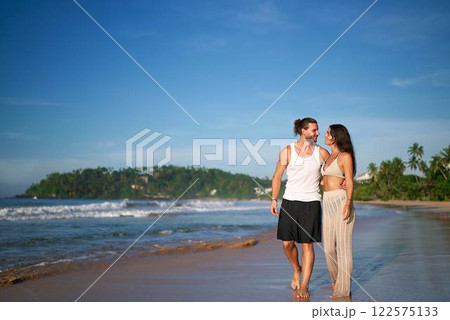 Romantic couple walks on sandy beach at sunset. Barefoot man, woman in casual summer attire hold hands, enjoying seaside honeymoon stroll. Tranquil ocean scenery, palm trees, vacation lovers leisure. Romantic couple walks on sandy beach at sunset. Barefoot man, woman in casual summer attire hold hands, enjoying seaside honeymoon stroll. Tranquil ocean scenery, palm trees, vacation lovers leisure. 122575133