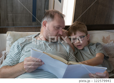Father hugs his little school-age son and reads funny paper literature. Happy little boy reading a book, enjoying his free weekend with his joyful father on the sofa Father hugs his little school-age son and reads funny paper literature. Happy little boy reading a book, enjoying his free weekend with his joyful father on the sofa 122579941