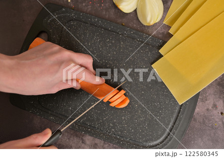 Woman's hand cutting carrot into slices on the black kitchen cutting board, top view. Lasagne pasta sheets, onion. Black background. Homemade lasagna cooking preparation Woman's hand cutting carrot into slices on the black kitchen cutting board, top view. Lasagne pasta sheets, onion. Black background. Homemade lasagna cooking preparation 122580345