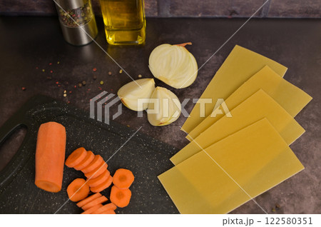 Carrot on the black kitchen cutting board. Lasagne pasta sheets, onion, oil, pepper. Black background. Homemade lasagna cooking preparation 122580351