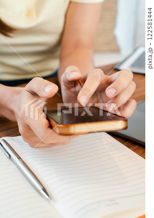 Hands holding a smartphone over a notebook with a pen on a wooden desk, capturing a moment of modern work or study. 122581444