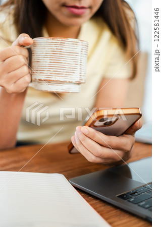 Close-up of a woman holding a coffee mug and smartphone, sitting at a desk with a laptop and notebook. 122581446