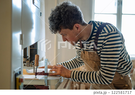 Skilled male carpenter concentrates on jigsaw machine in shop, carefully shaping small wood parts 122581717