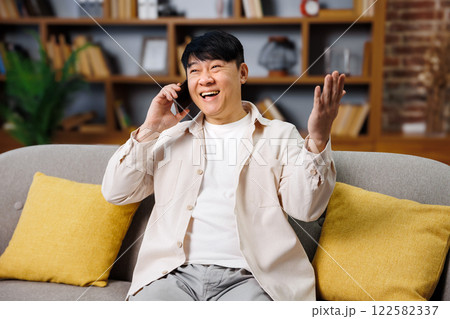 A man expresses joy and laughter while speaking on the phone in a relaxed living space. Bright yellow cushions add warmth to the cozy atmosphere, enhancing the friendly interaction. 122582337