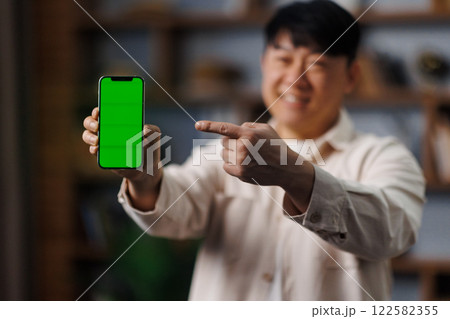Individual is happily showcasing a smartphone with a vibrant green display in a warm, inviting room filled with personal items. The gesture highlights the device's screen. Individual is happily showcasing a smartphone with a vibrant green display in a warm, inviting room filled with personal items. The gesture highlights the device's screen. 122582355