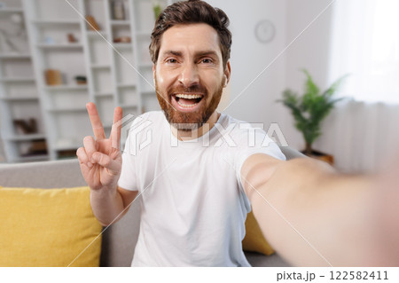 takes selfie, video call, shows peace symbol, cheerful young man takes a selfie in his cozy living room. He flashes a peace sign with one hand while smiling brightly, surrounded by yellow cushions and 122582411