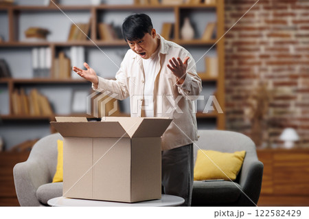 opening delivery box, excited man, Online shopping concept, man stands surprised as he opens a large cardboard box in a cozy living room. The setting features a well-organized bookshelf and 122582429