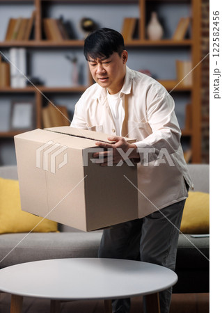 opening delivery box, excited man, Online shopping concept, man is carefully lifting a large cardboard box in a warmly decorated living room. The space features a comfortable sofa and stylish shelves opening delivery box, excited man, Online shopping concept, man is carefully lifting a large cardboard box in a warmly decorated living room. The space features a comfortable sofa and stylish shelves 122582456